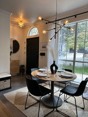 A dining area with a round table set for four, black chairs, a modern chandelier, and a view through large windows.