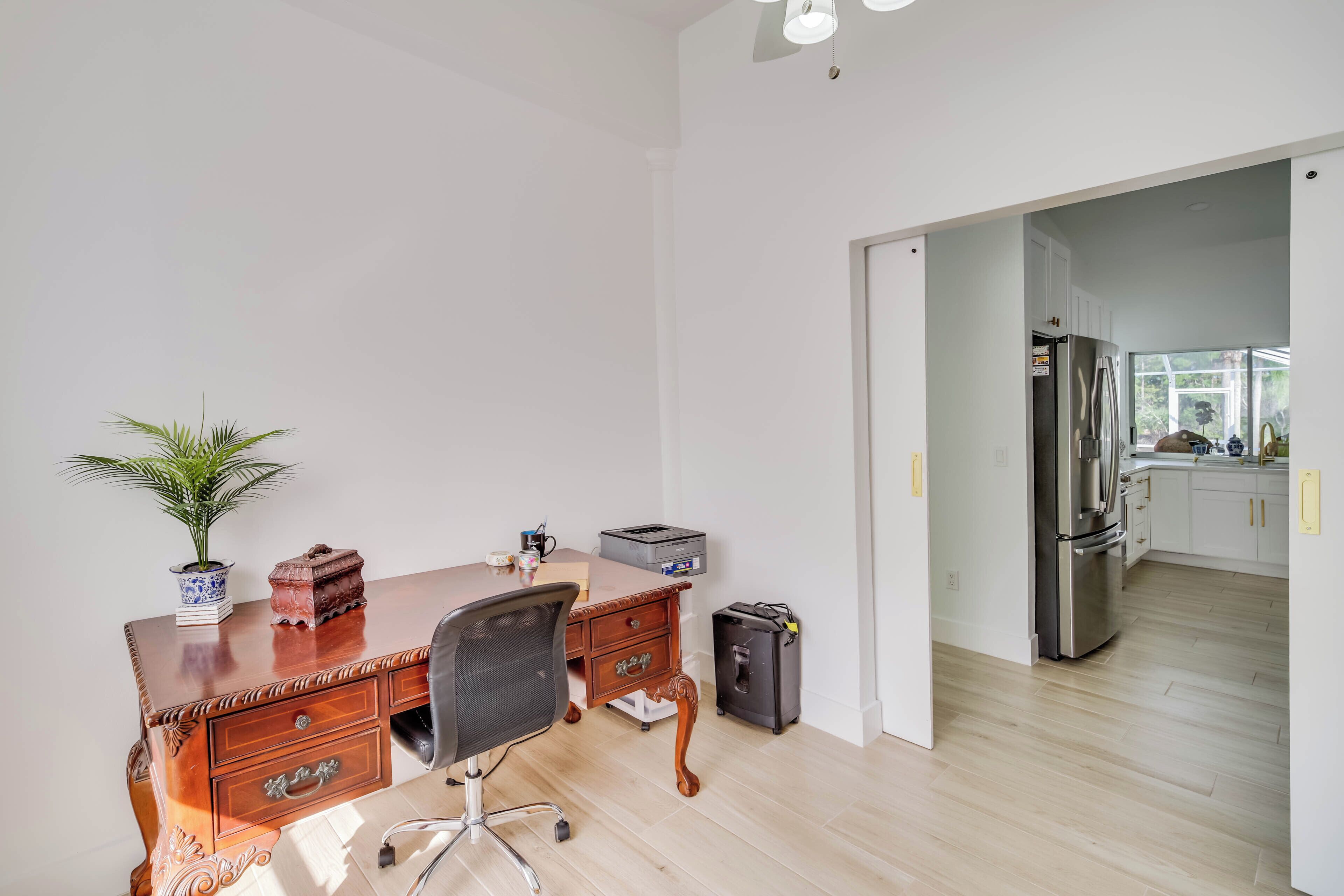 A room with a wooden desk, a chair, a small potted plant, a trash bin, and a refrigerator in the background.