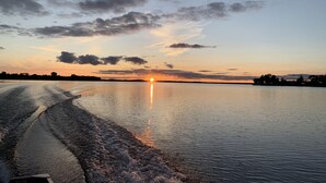 A calm lake at sunset with a boat wake, trees in the distance, and a clear sky.