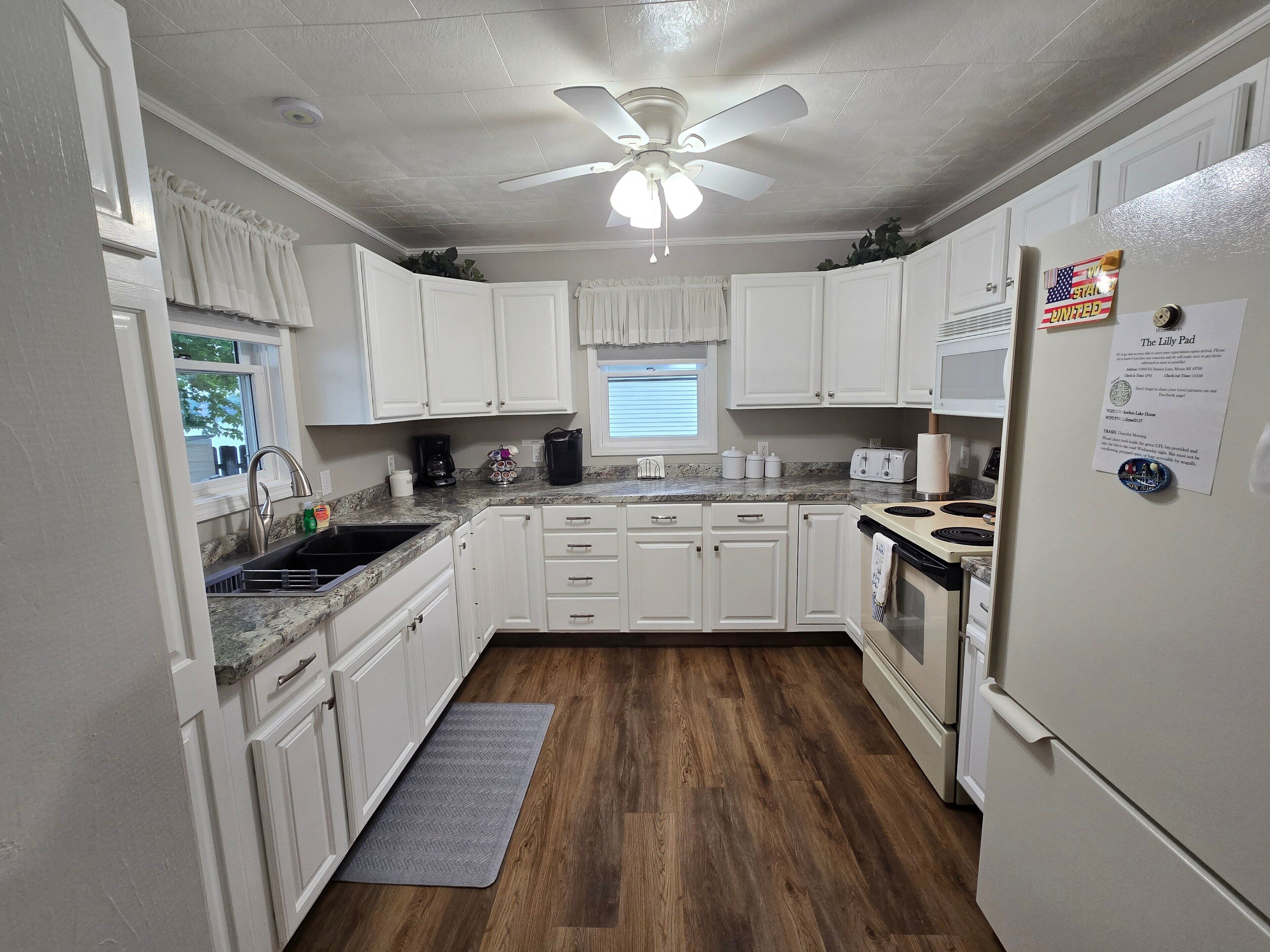 Une cuisine avec des armoires blanches, un comptoir en granit, un ventilateur de plafond, un réfrigérateur et un plancher en bois.