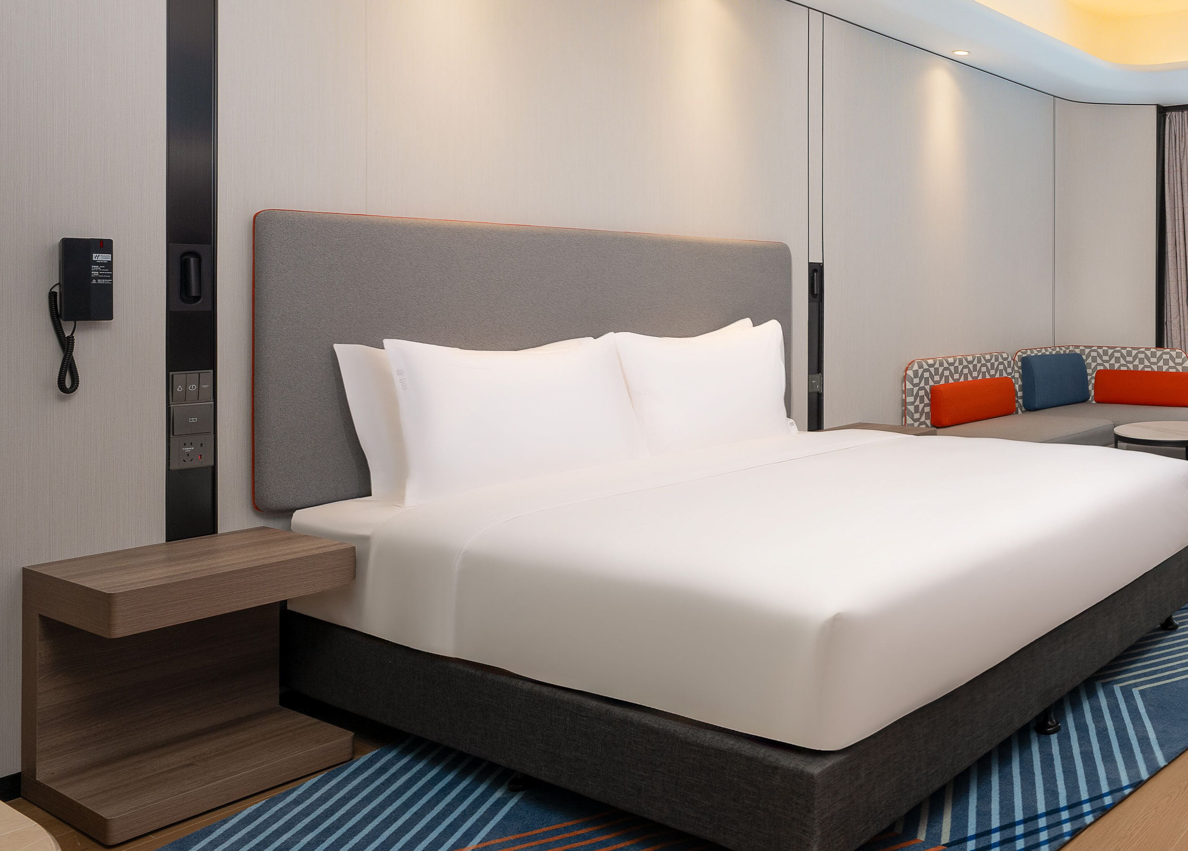 Une chambre d’hôtel moderne dotée d’un grand lit, d’une table de chevet, d’un téléphone intégré et d’un coin salon agrémenté de coussins colorés.