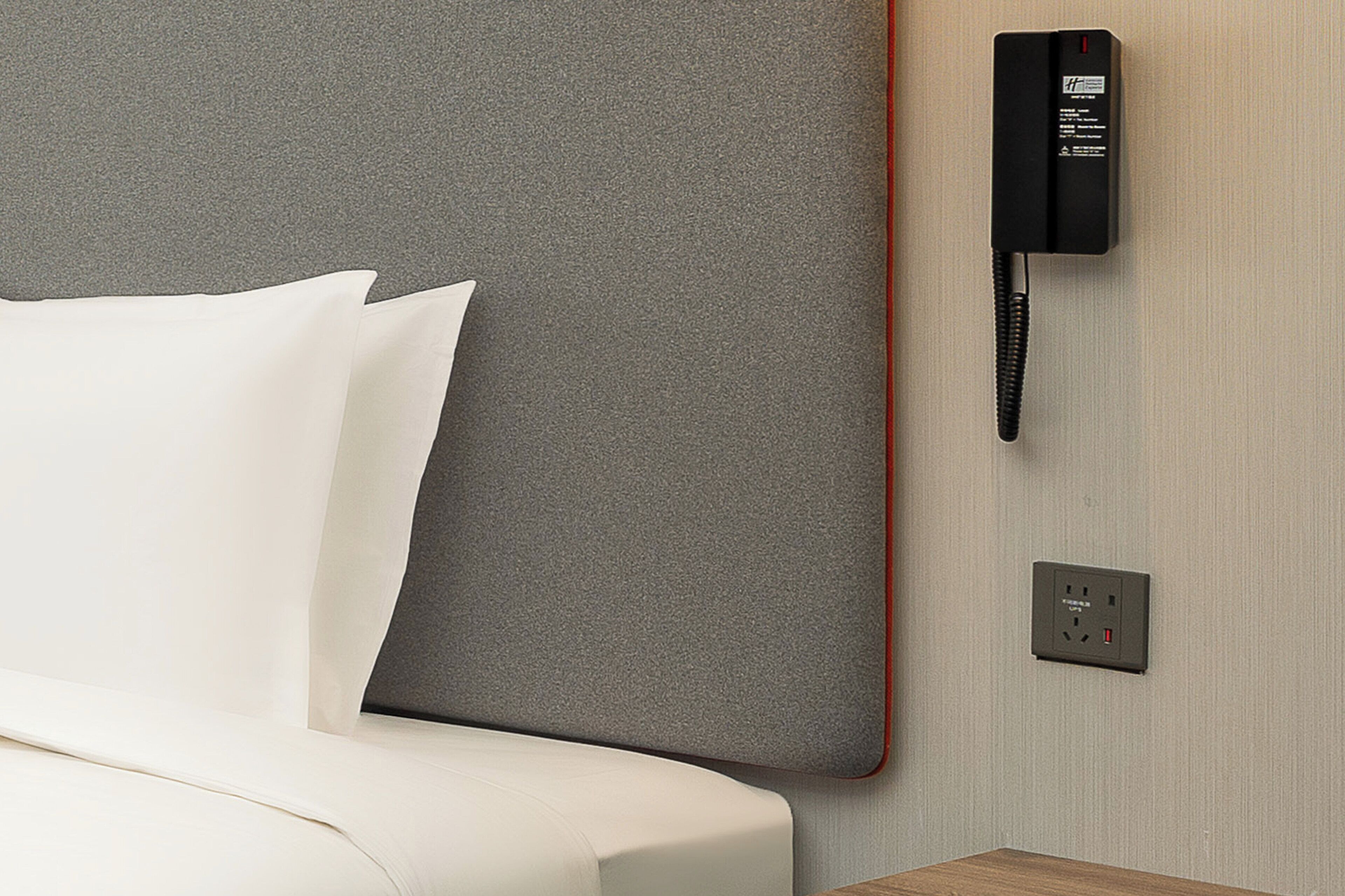 Habitación de hotel con cabecera gris, cama blanca, teléfono negro y toma de corriente en la pared.