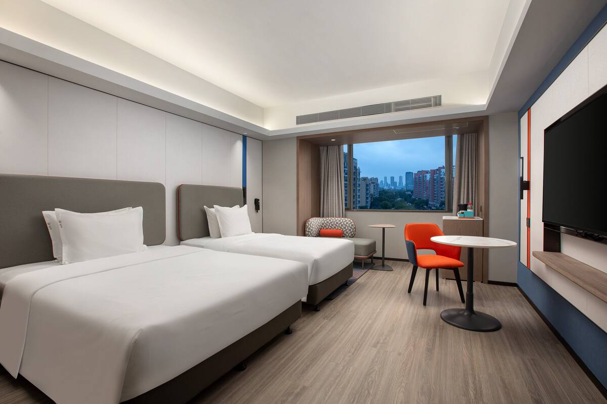 A hotel room with two beds, a television, a small table, and a window with a city view.