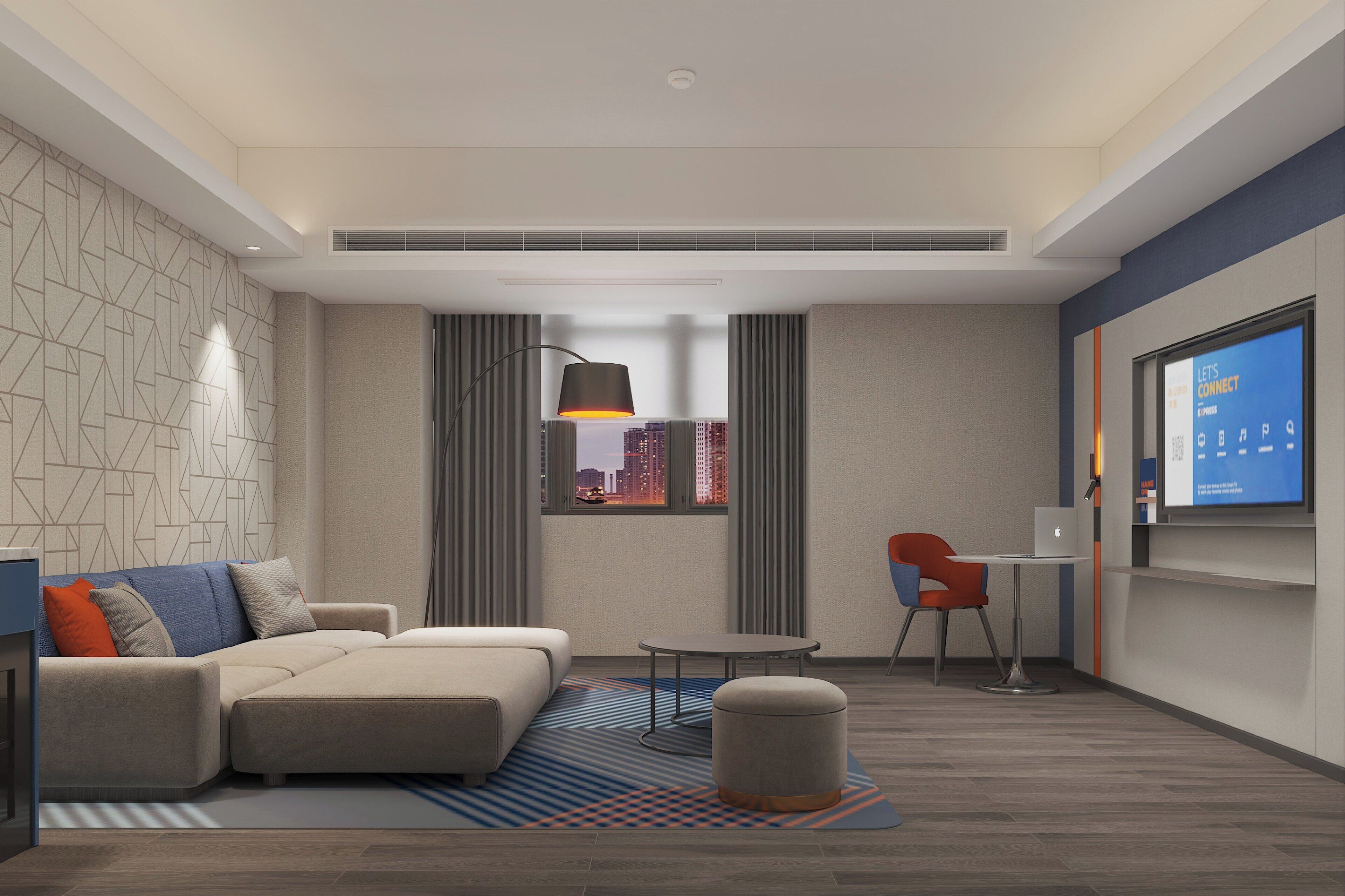Een moderne hotelkamer met een hoekbank, een tafeltje, een rode stoel, een rond poef, uitzicht op de stad door het raam, en een digitaal scherm waarop staat 'Laten we verbinden'.
