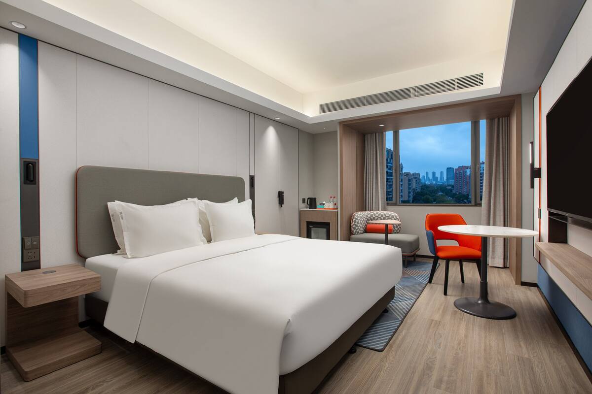 A modern hotel room with a large bed, a desk, a chair, and a city view.