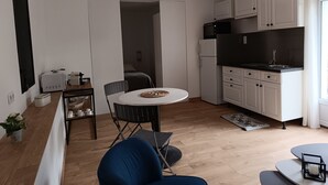 A modern kitchen with white cabinets, a black microwave, and a refrigerator. There is a dining area with a round table and chairs, and a small kitchenette with a sink and stove. - Rez de Villa au Calme T2 + Terrasse Privative sur la Colline (Vidauban)