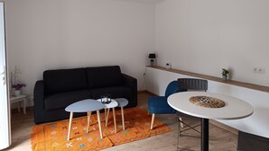 A modern living room with a black sofa, a small white table, a blue chair, and a round table with a woven placemat. - Rez de Villa au Calme T2 + Terrasse Privative sur la Colline (Vidauban)