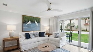 Een moderne woonkamer met een witte modulaire bank, twee bijzettafels met lampen, een ronde salontafel, een groot schilderij van vogels, en uitzicht op een groen gazon en palmbomen door de glazen deur.