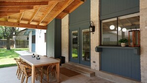 Eine Terrasse im Freien mit Holztisch und -stühlen, ein überdachter Bereich mit Holzboden und ein großes Fenster mit Blick auf den Garten.
