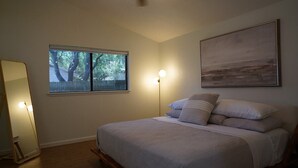 Ein Schlafzimmer mit einem Bett, einem Fenster mit Blick auf Bäume, einem Bild an der Wand und einer Stehlampe.