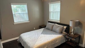Un dormitorio con cama, dos ventanas, una mesita de noche y una lámpara.