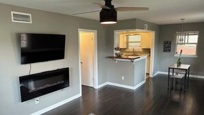 Una sala de estar moderna con un televisor de pantalla plana, estanterías empotradas, un ventilador de techo y una cocina al fondo.