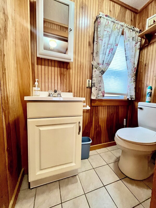 Un baño pequeño con paneles de madera, un lavamanos blanco con espejo, un inodoro y una ventana con cortinas estampadas.