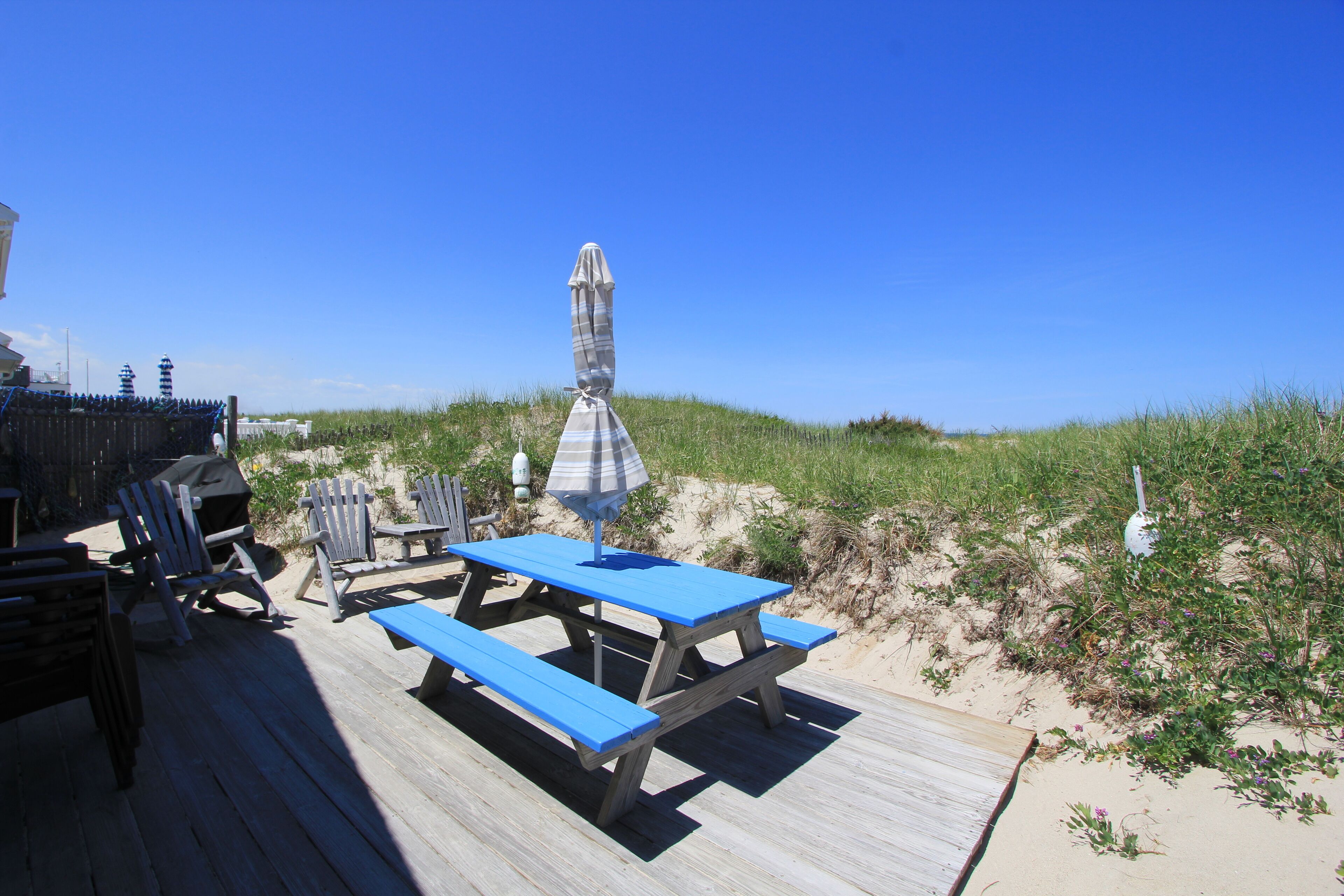 A wooden deck with picnic tables and chairs, overlooking a beach with dunes.