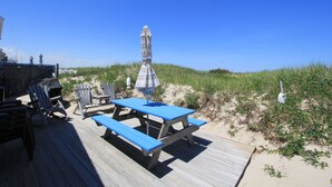Une terrasse en bois, dotée de tables et de chaises pour pique-niquer, qui offre une vue sur une plage bordée de dunes.