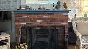 Une cheminée avec un foyer en briques, une étagère ornée d’objets décoratifs et un téléviseur posé sur un meuble, à gauche.