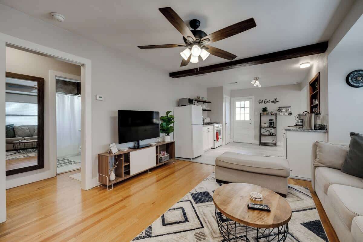 A modern living room with a flat-screen TV, a ceiling fan, a dining area, and a kitchen in the background.