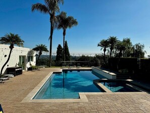 Una piscina rectangular con una solana con baldosas, rodeada de palmeras y un edificio blanco con una puerta y ventanas.