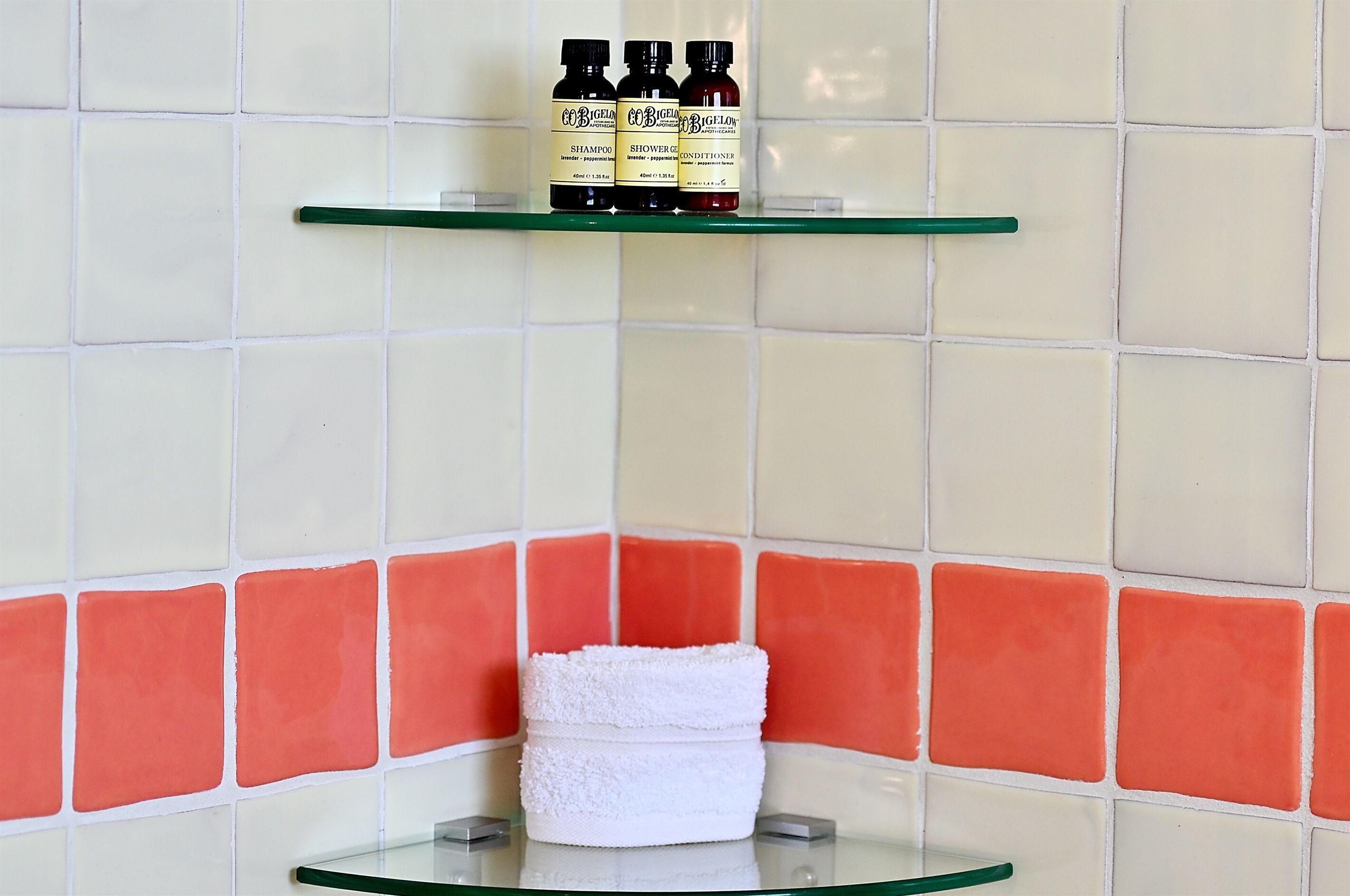 A bathroom with a glass shelf holding three bottles of shampoo, conditioner, and shower gel. A white towel is placed on a glass surface below the shelf.