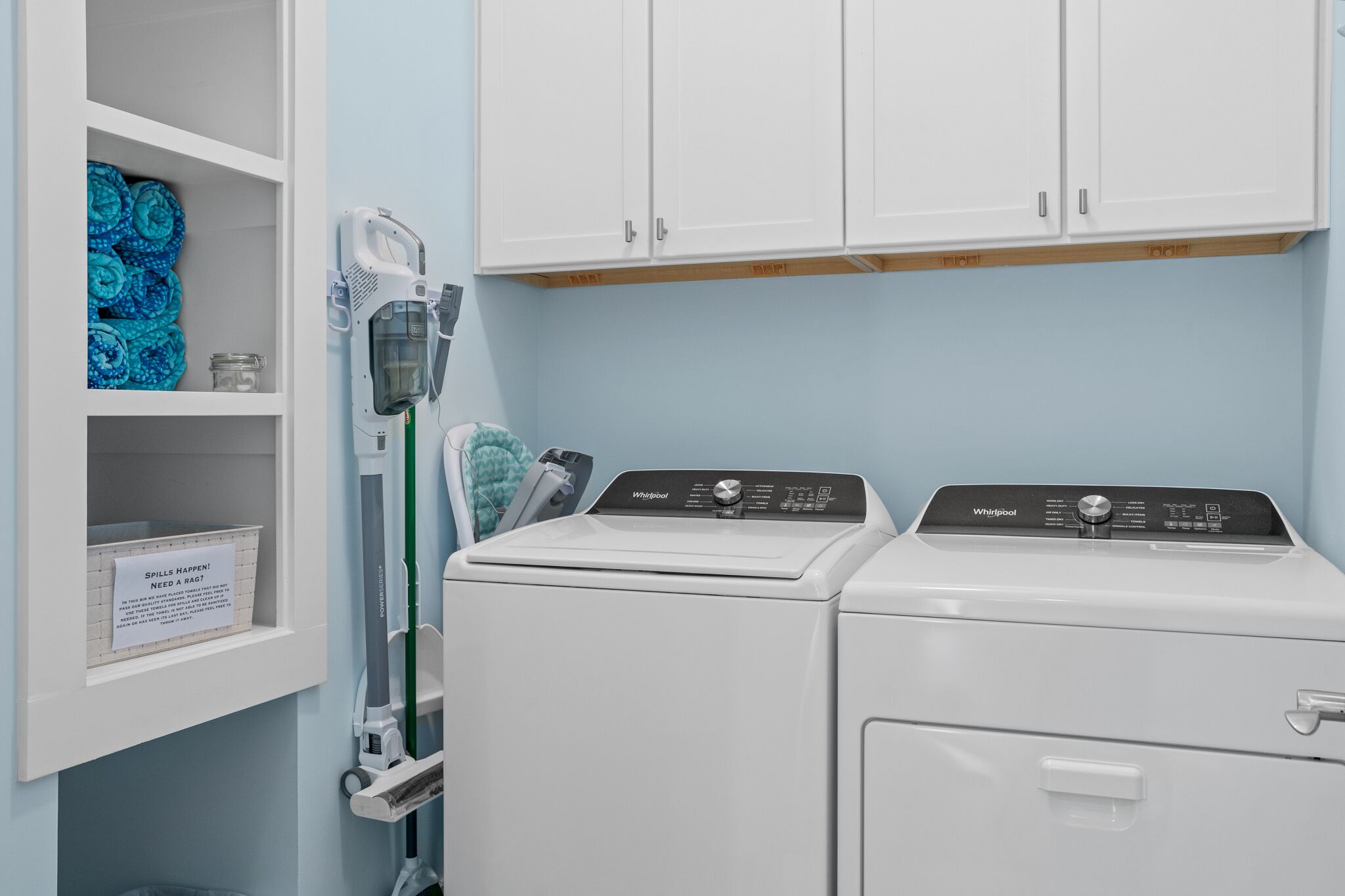 Une buanderie équipée de deux lave-linge Whirlpool blancs, d’une étagère avec des serviettes pliées et d’un panneau indiquant la procédure à suivre en cas de déversement de liquide.