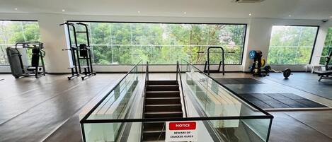 An indoor gym with exercise equipment, a staircase with a glass barrier, and large windows offering a view of greenery.