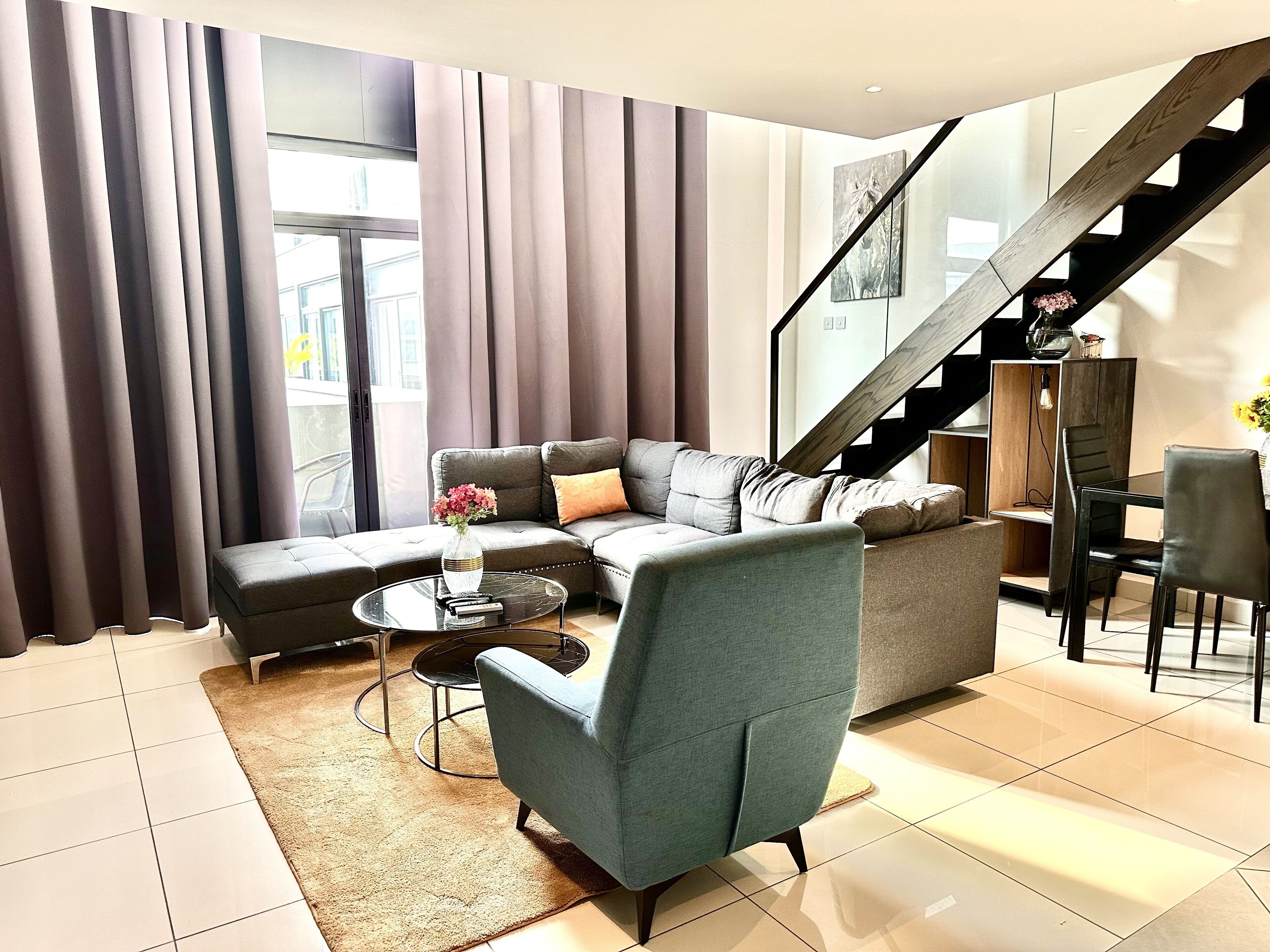 A modern living room with a sectional sofa, a glass coffee table, and a grey armchair. A staircase is visible in the background.
