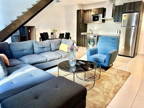 A modern living room with a grey sectional sofa, a blue armchair, and a coffee table. - Embassy Gardens Apartment (Accra)