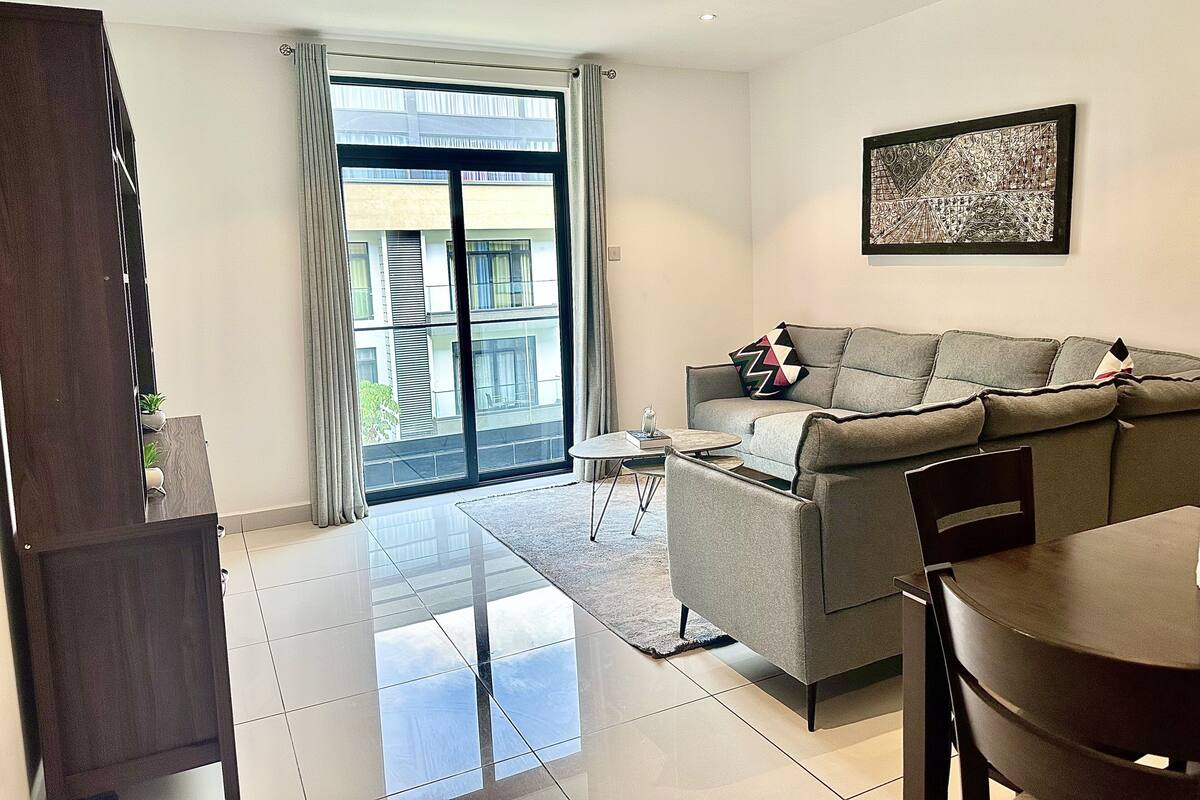 A modern living room with a grey sofa, a dining table, and a large window with a view of buildings.