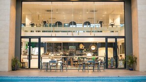 A modern hotel with a glass facade, a pool area in front, and a dining area with outdoor seating. - Embassy Gardens Apartment (Accra)