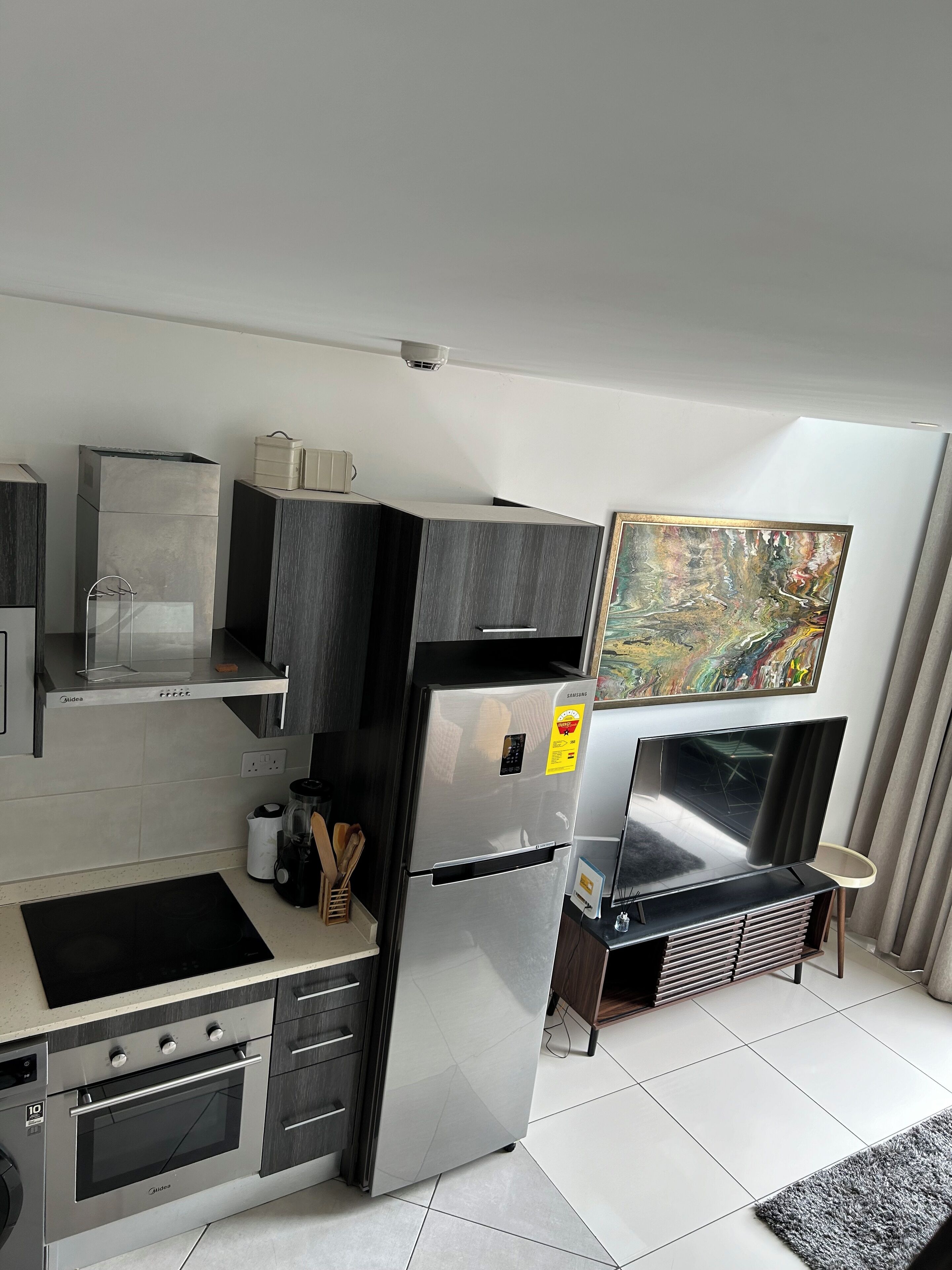 Una cocina moderna con horno empotrado, refrigerador de acero inoxidable y microondas. Hay un cuadro en la pared y una pequeña mesa con una silla.