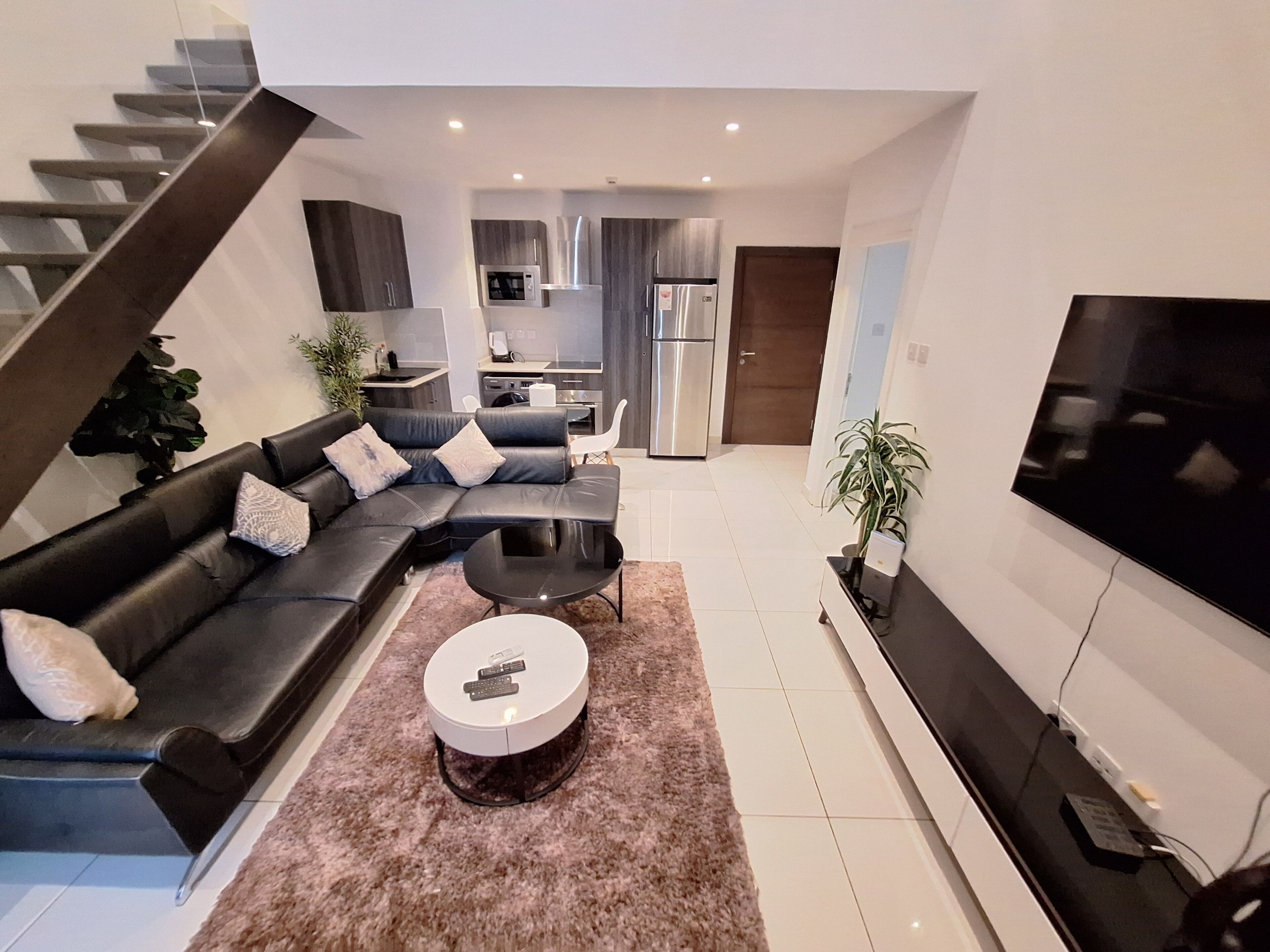 A modern living room with a sectional sofa, a coffee table, and a TV. There is a kitchen in the background with a refrigerator and microwave.