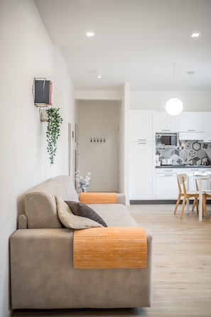 A modern living room with a sofa, a dining area with a table and chairs, and a kitchen with white cabinets and a microwave.
