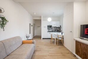 A modern living room with a sofa, a dining area with a table and chairs, a kitchen with white cabinets, and a TV on a wooden cabinet.