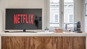 Eine moderne Küche mit einem großen Flachbildfernseher, der das Netflix-Logo zeigt, einer Kaffeemaschine, einer Teebox und einem Fenster mit Blick auf Gebäude.
