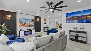 A modern living room with a grey sofa, a ceiling fan, and a cityscape painting on the wall. - Hot tub, Gazebo, Firepit Table Getaway Oasis House!!!! (Fort Myers)