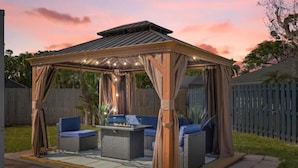 A covered outdoor seating area with a fire pit, cushioned chairs, and a pergola structure. - Hot tub, Gazebo, Firepit Table Getaway Oasis House!!!! (Fort Myers)
