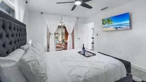 A bedroom with a bed, a ceiling fan, a painting of a beach, and a window with curtains. - Hot tub, Gazebo, Firepit Table Getaway Oasis House!!!! (Fort Myers)