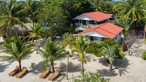 A beachfront property with a two-story building, palm trees, and wooden lounge chairs on the sand. - Complete Beach Retreat: Cottage + Beachhouse (Placencia)
