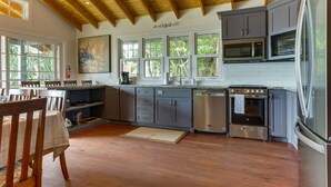A modern kitchen with wooden beams, grey cabinets, stainless steel appliances, and a large window overlooking greenery. - Complete Beach Retreat: Cottage + Beachhouse (Placencia)