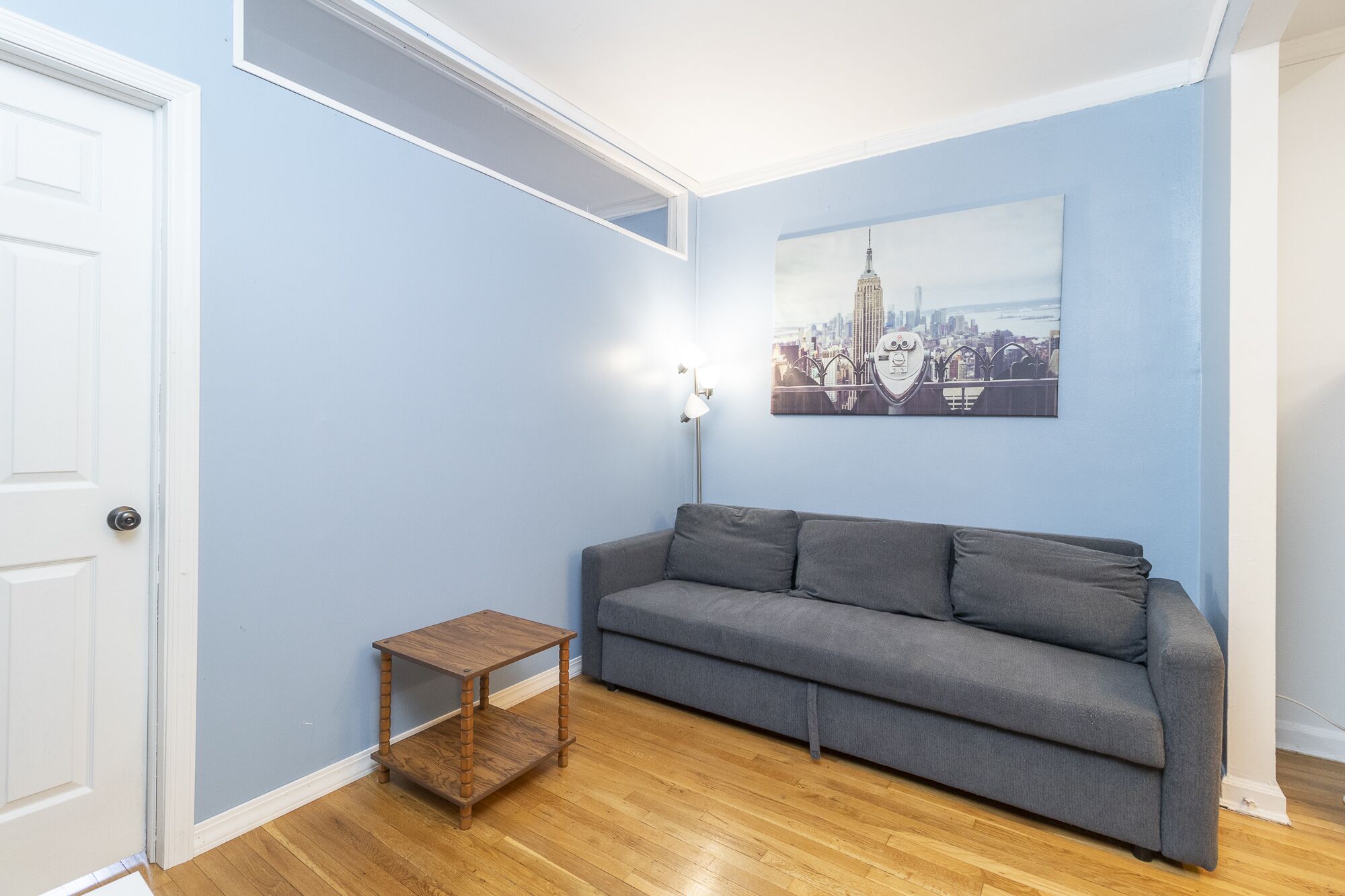 A room with a grey sofa, a small wooden side table, a floor lamp, and a cityscape painting on the wall.