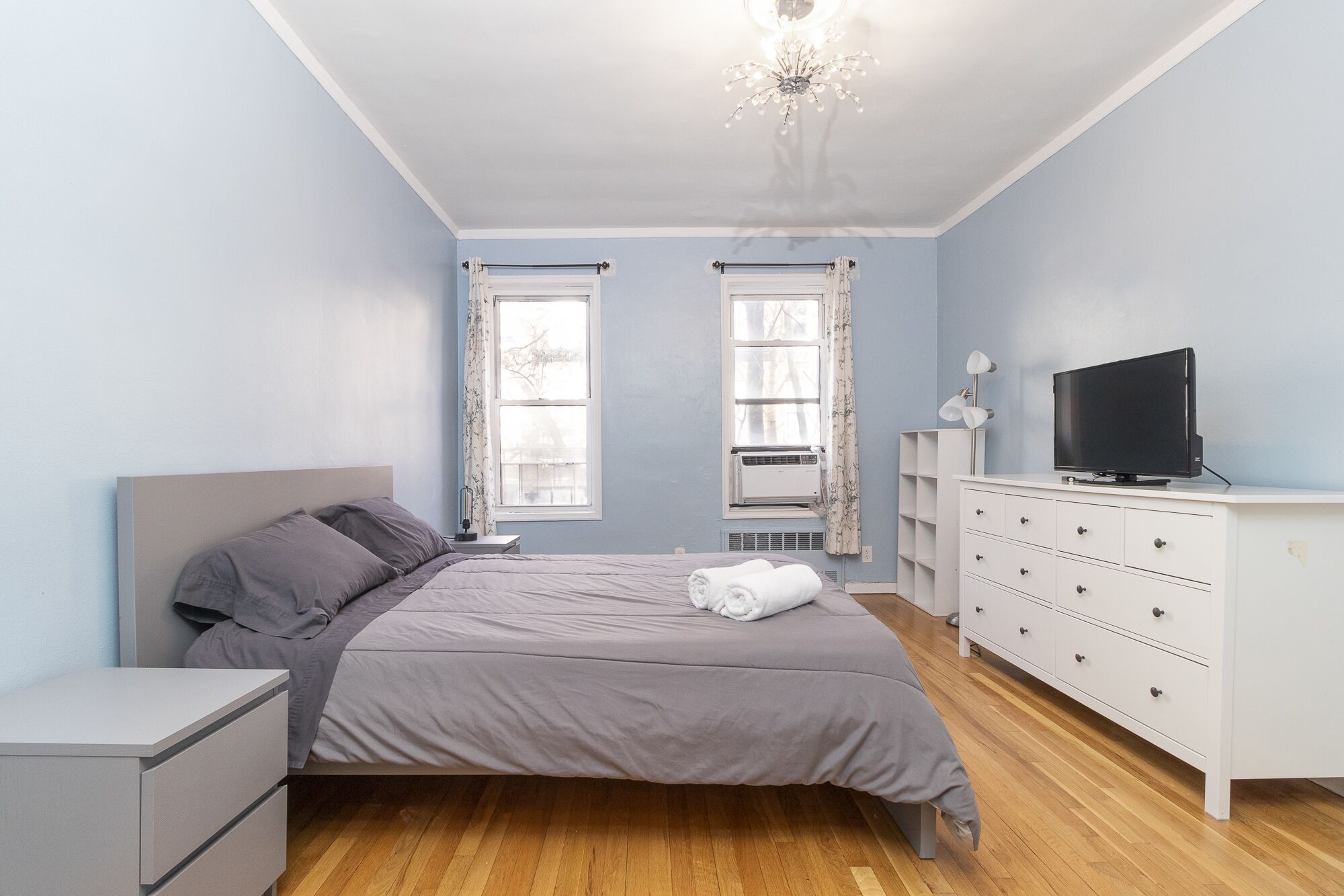 Un dormitorio con cama, cómoda, televisor y dos ventanas con cortinas.