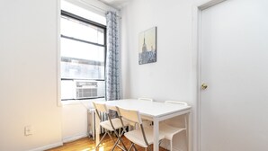 Een lichte, kleine kamer met een witte tafel en stoelen, een raam met gordijnen en een radiator.