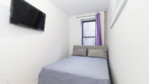 A small, neatly arranged bedroom with a bed, a window with purple curtains, and a mounted television.