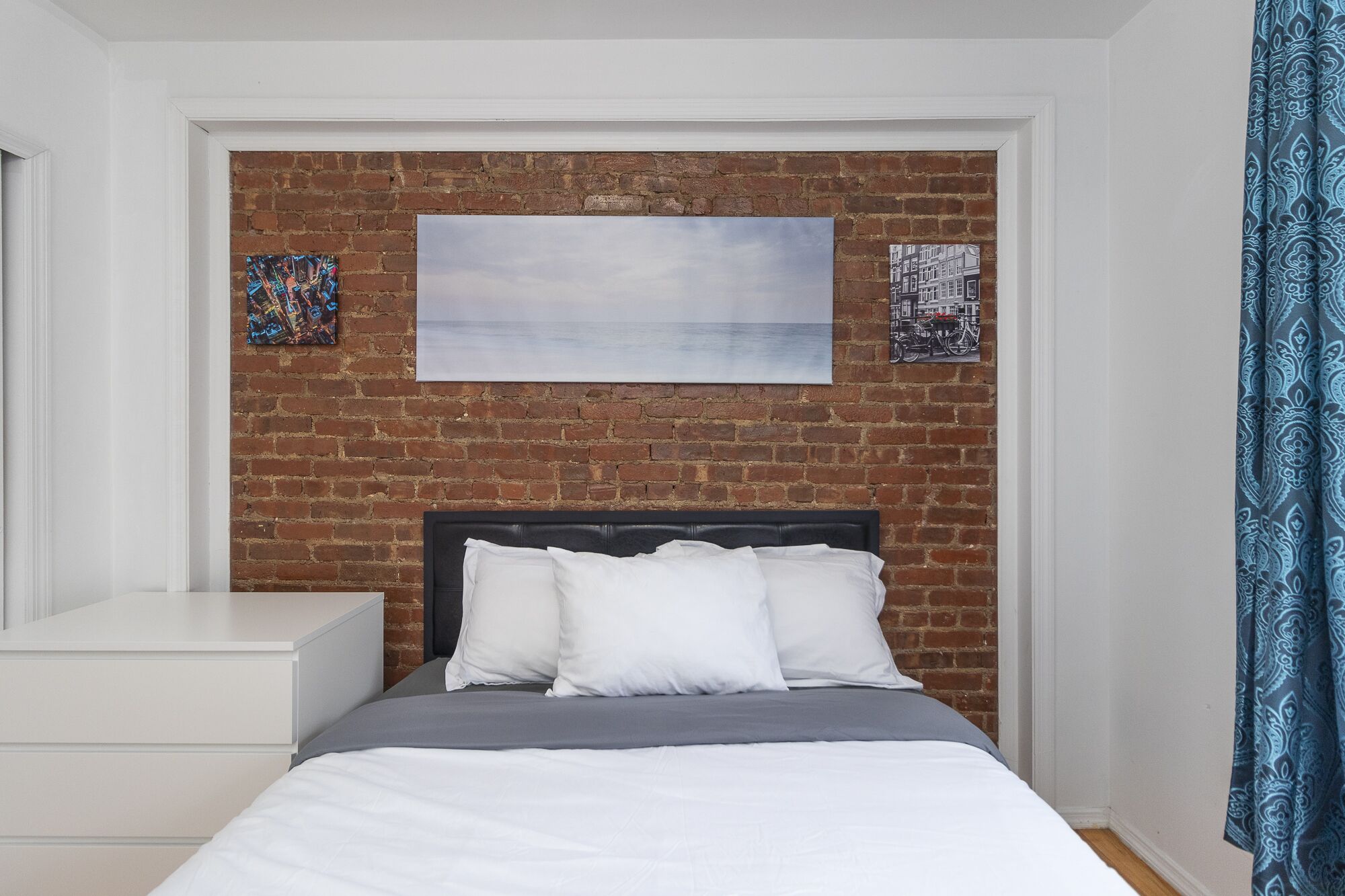 Un dormitorio con pared de ladrillo, una cama con ropa de cama blanca, una mesita de noche blanca y un cuadro enmarcado sobre la cama.