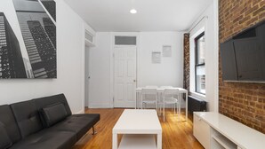 A modern living room with a black sofa, a white coffee table, and a TV mounted on a brick wall. There are two white chairs and a white table in the background. - Upper East Modern & Chic Two BR Apt near Central Park (New York)