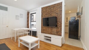 A compact living space with a white table and chairs, a brick accent wall, a flat-screen TV, and a black refrigerator. - Upper East Modern & Chic Two BR Apt near Central Park (New York)