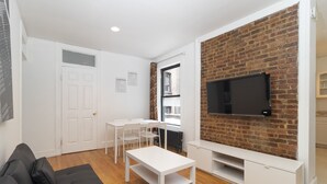 A modern living room with a brick accent wall, a flat-screen TV, a white sofa, a dining table with chairs, and a white cabinet. - Upper East Modern & Chic Two BR Apt near Central Park (New York)