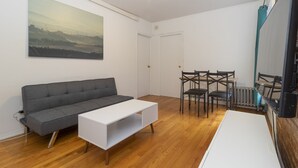 A modern living room with a grey sofa, white coffee table, and a dining area with four chairs. A large landscape painting is on the wall, and there is a white door and radiator visible. - Spacious Two BR Apartments in Yorkville Upper East NYC (Manhattan)