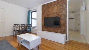 A modern living room with a brick accent wall, a flat-screen TV mounted above a white entertainment unit, a wooden dining table with chairs, and a grey sofa. - Spacious Two BR Apartments in Yorkville Upper East NYC (Manhattan)