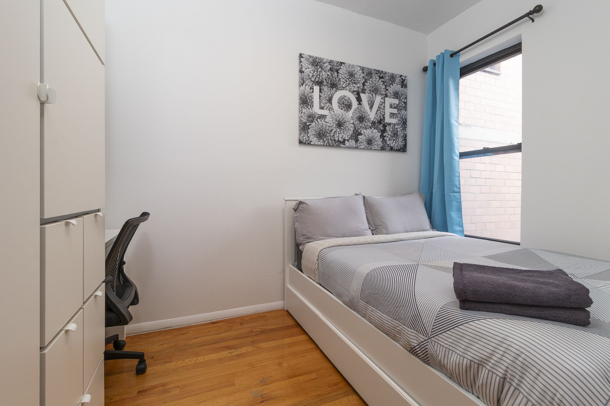 A bedroom with a bed, a desk, and a chair. There is a window with blue curtains and a picture on the wall that says "LOVE".